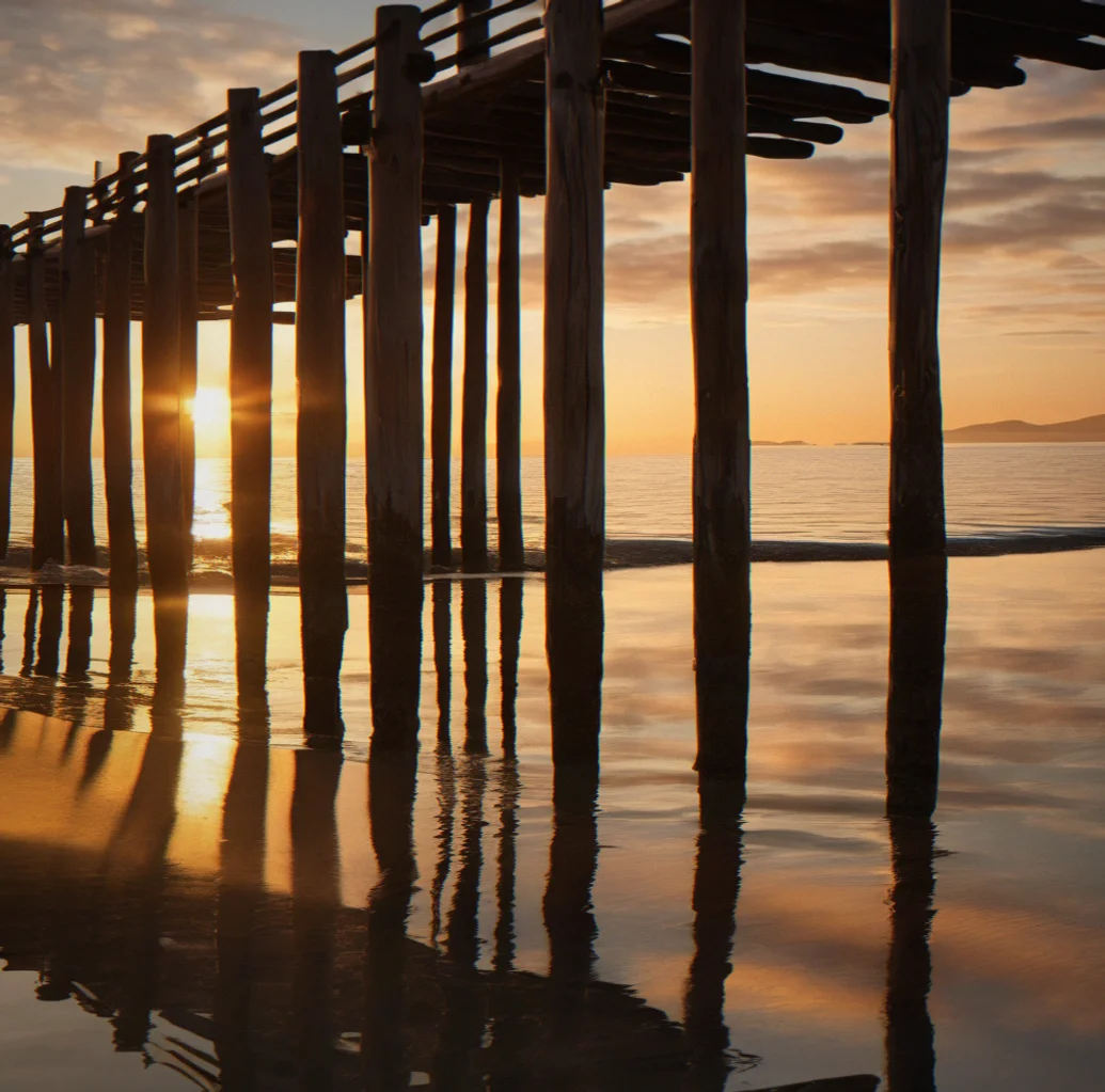 Sunrise light breaking across calm bay with wooden pier pilings casting long shadows