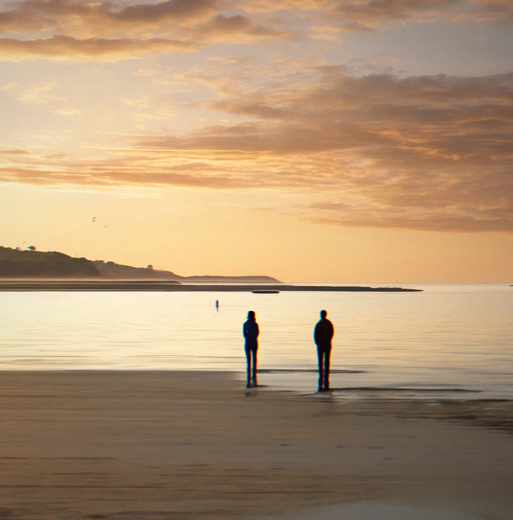 Sunset view across estuary with two people standing at water's edge, neither looking at camera, arms at rest
